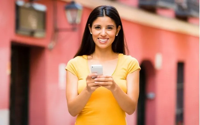Latina woman with a hairstyle with a part down the middle holds a phone while standing in front of a brightly-colored marketplace