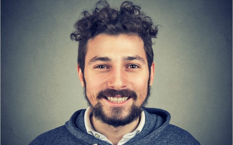 Man with an unkept beard and a curly top bowl cut smiles big at the camera against a gradient gray background
