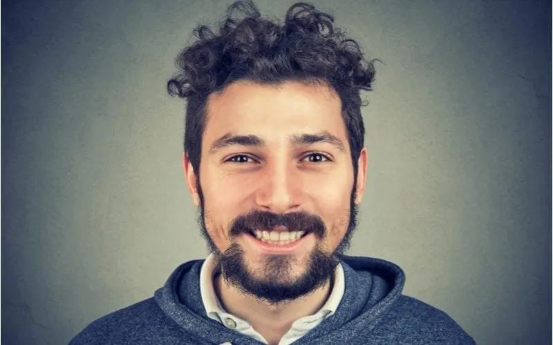 Man with an unkept beard and a curly top bowl cut smiles big at the camera against a gradient gray background