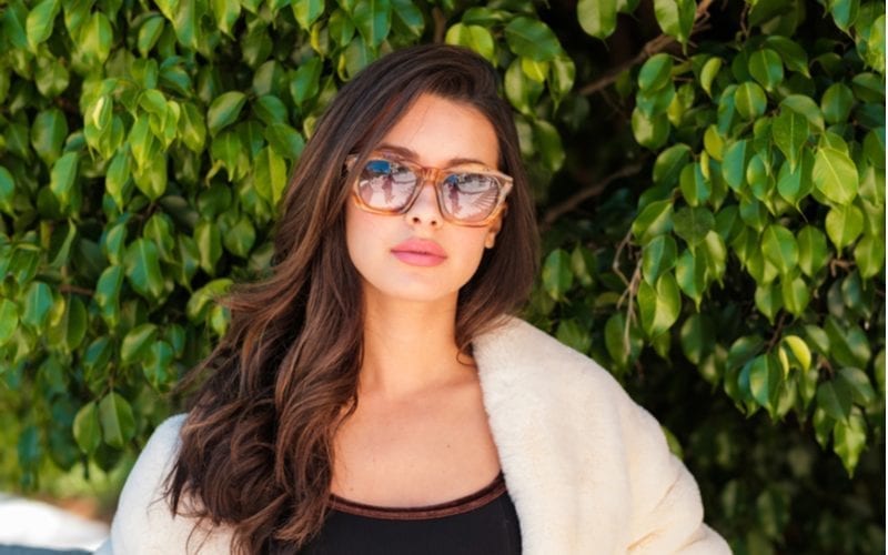 Young latina woman with long hair in sunglasses stands in front of a bush