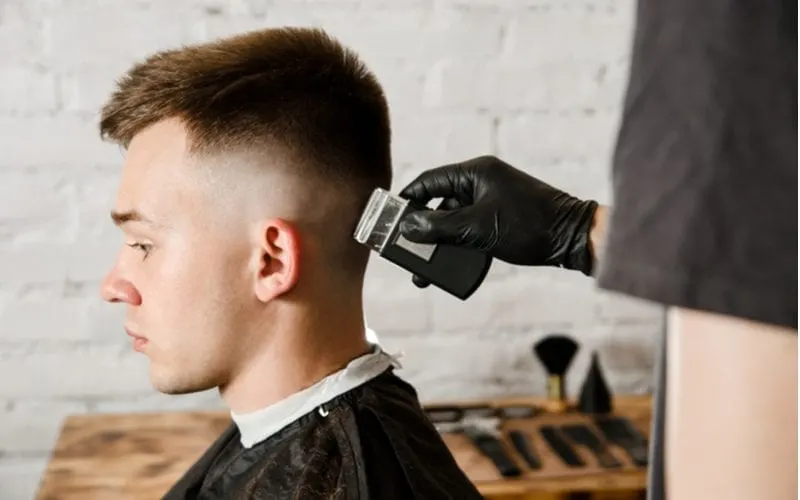 Guy with a tapered fade gets the back of his neck styled with help from a barber in a black shirt and gloves
