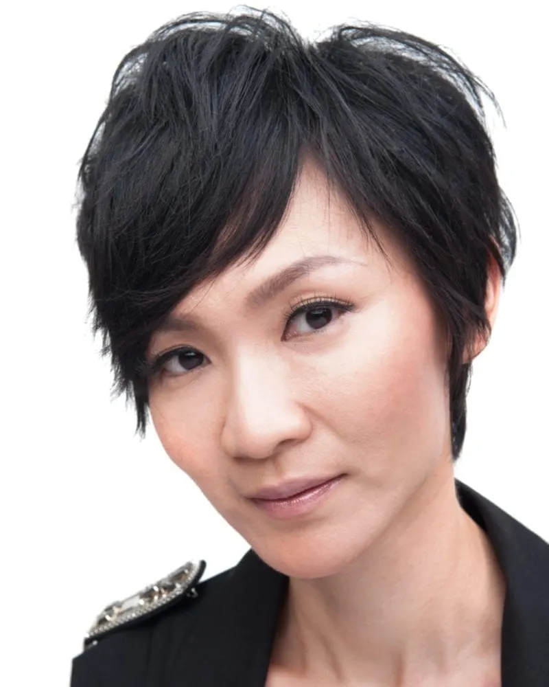 Asian woman with short hair and a black blazer with studded shoulder pads looks at the camera without smiling