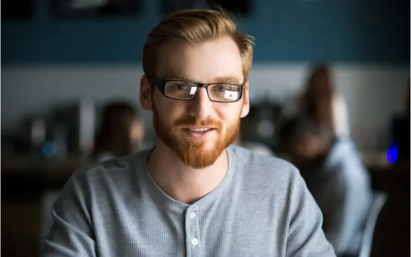 Man with glasses sits in a cafeteria wearing a mens redheaded hairstyle and grey shirt
