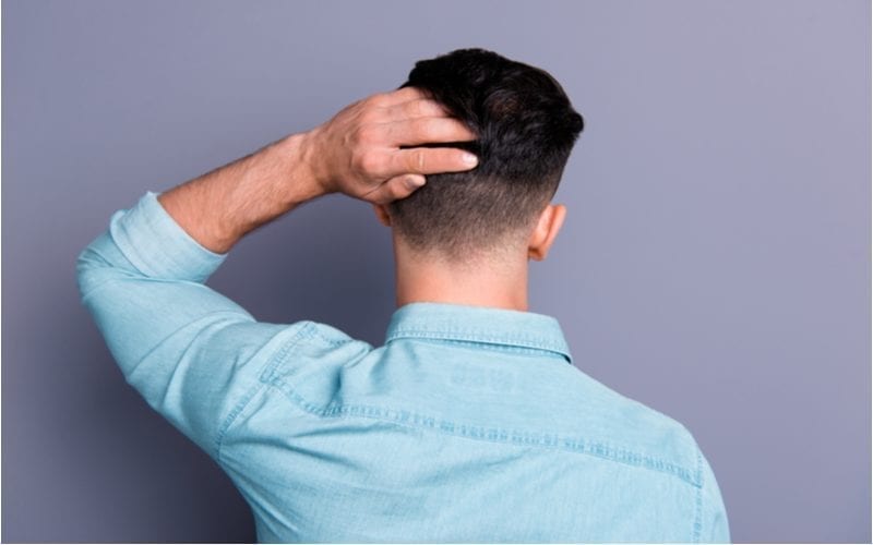Guy in a blue denim button-up shirt stands against a purple wall and holds his taper fade haircut