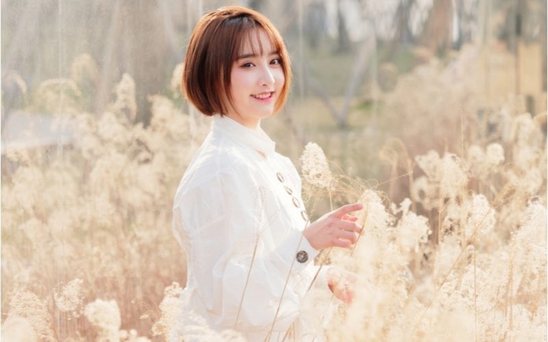 Short-haired asian woman stands in a field of white flowers while wearing a white dress