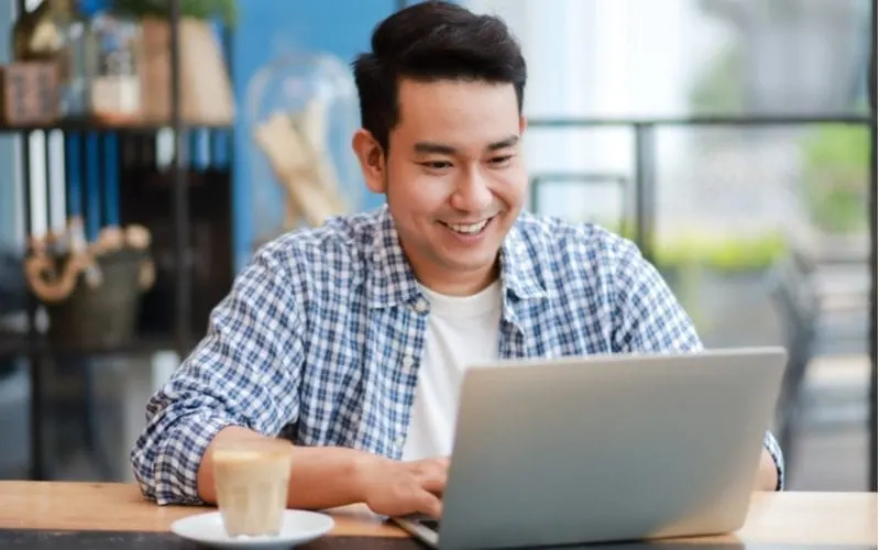 Asian man in an unbuttoned long sleeve shirt smiling and looking at a laptop