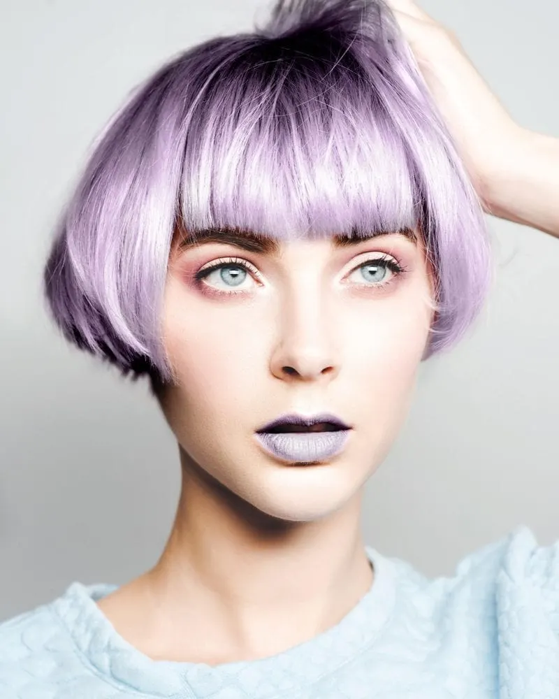 Woman with lilac hair and a bowl cut holds the top of her hair and wears lilac lipstick