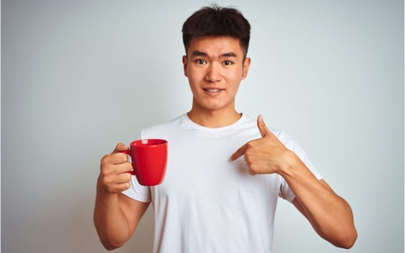Chinese man holding a red mug and pointing to himself while wearing a white tshirt and a rocking a bowl haircut in a studio