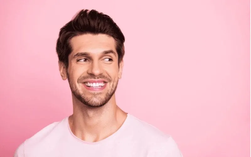 Against a pink background is a man with a tapered fade wearing a white crew neck shirt and smiling while looking to his right
