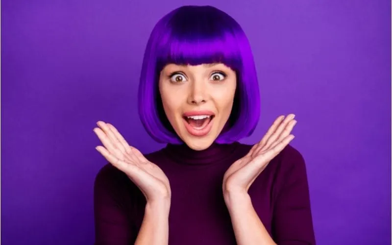Woman with purple hair holds her hands up to her face in exclamation and excitement
