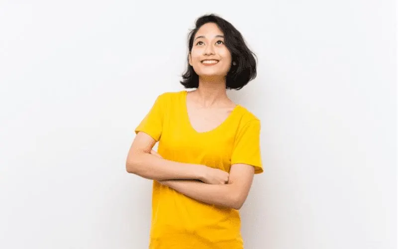 Asian woman with short hair and a yellow shirt crosses her arms and looks upward while smiling