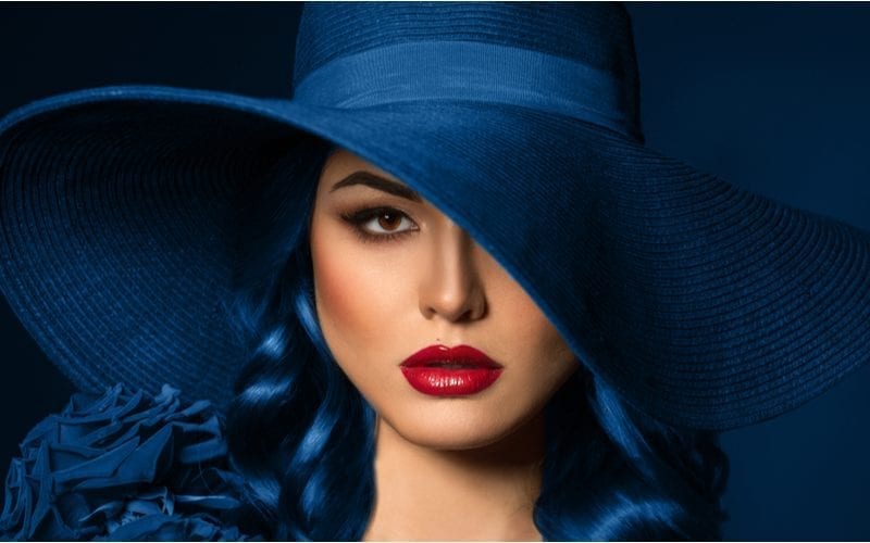 Woman with dark blue hair and a dark blue hat with bright red lipstick stands against a blue background