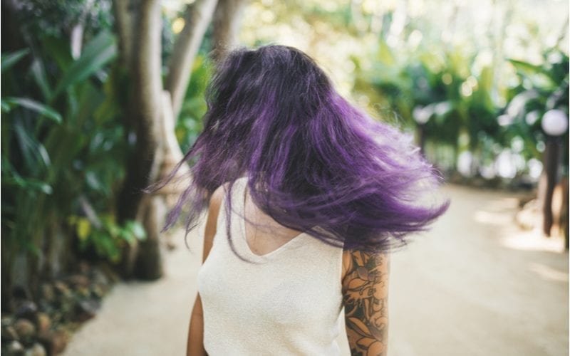 Woman with purple hair and a white semi-transparent shirt with a tattoo sleeve stands in a forest