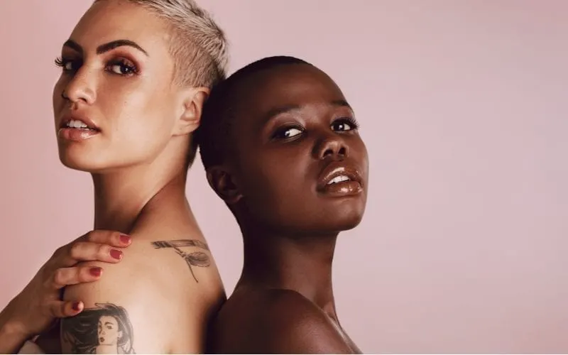 Two confident and beautiful women standing back to back with shaved heads in a studio with a pink backdrop