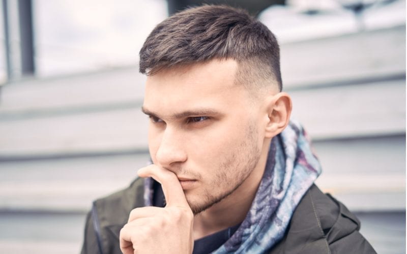 Man with a caesar tapered fade haircut holds his left finger up to his nose and thinks about something while sitting on steps