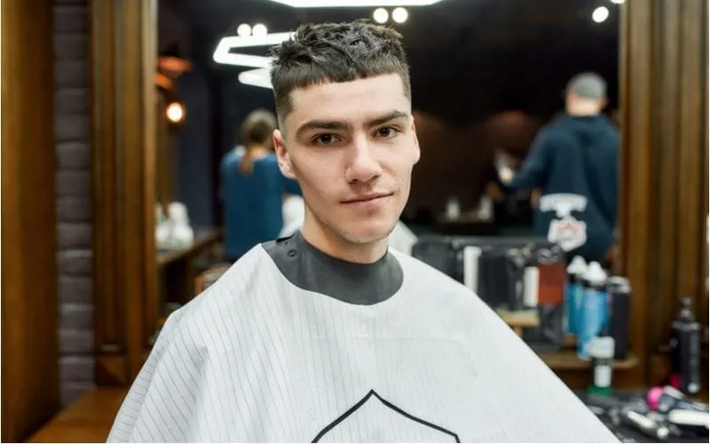 Man in a barbershop with a Caesar haircut sits and wears an apron