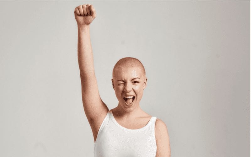 Woman with a shaved haircut holds up a fist and wears a tight white tank top in a studio