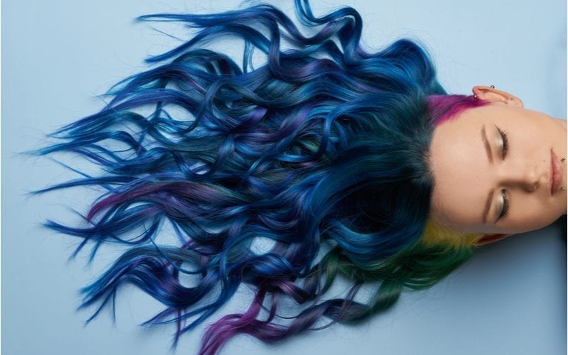 Lady with dark blue hair lays against a blue wall and has purple and yellow roots