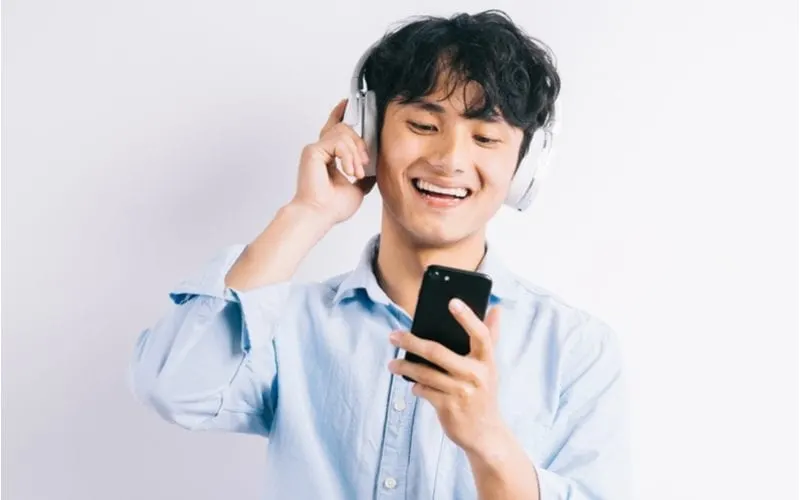 Young asian man smiling and looking at his phone and holding headphones that rest on top of his long bowl cut
