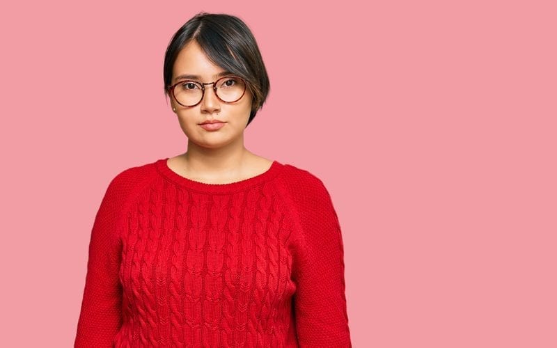 Pretty young woman with a Latina hairstyle wears a red sweater and round glasses while standing in a studio with a pink backdrop