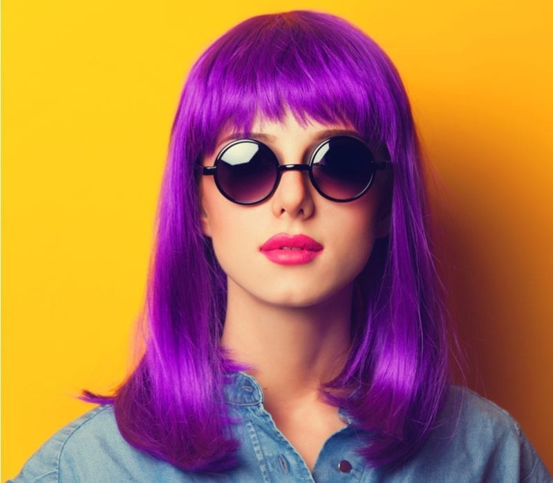 In a studio with an orange background, a woman with purple hair wears dark round sunglasses