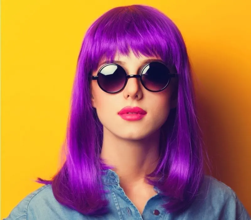 In a studio with an orange background, a woman with purple hair wears dark round sunglasses