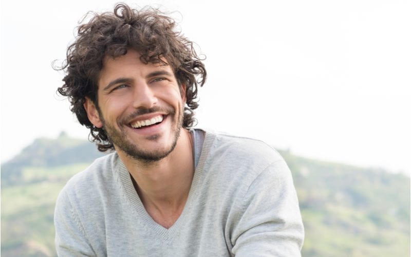 Guy with long curly hair and a grey sweater smiles and looks upward