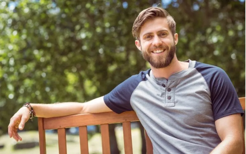 Young man relaxed with his arm on the top of a park bench while wearing a baseball shirt