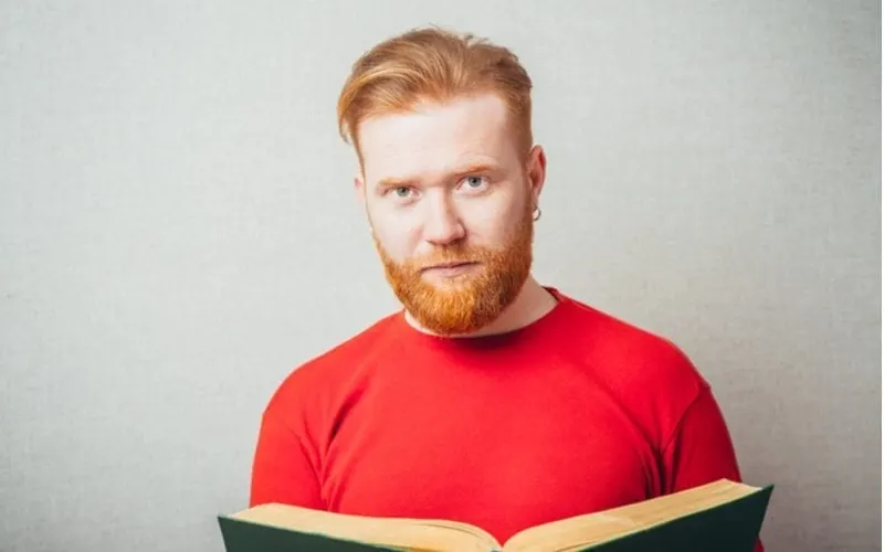 Man with a red shirt and a red headed hairstyle reads a book and stands against a grey background