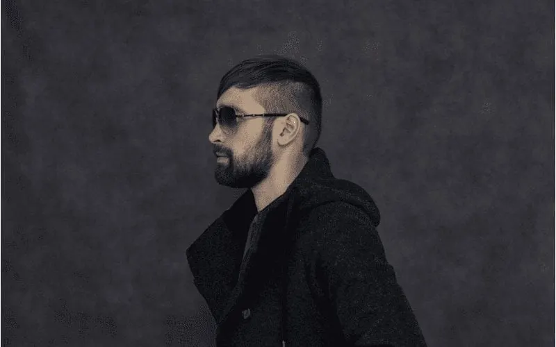 Man with a fade haircut and long top in sunglasses wearing black in a black room