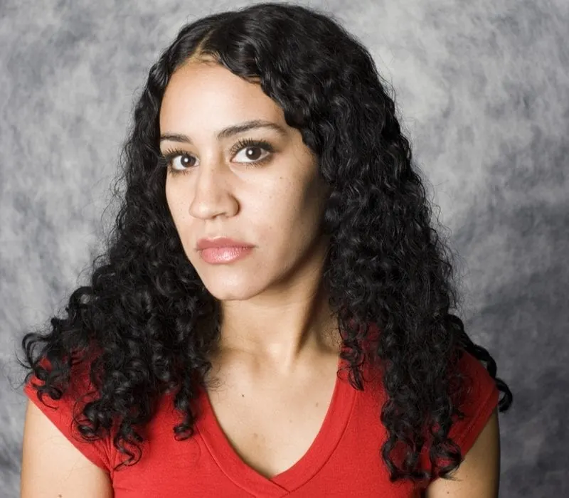 Latina woman with cascading curls (a typical Latina hairstyle) in a red v-neck stands in front of a marbled studio backdrop