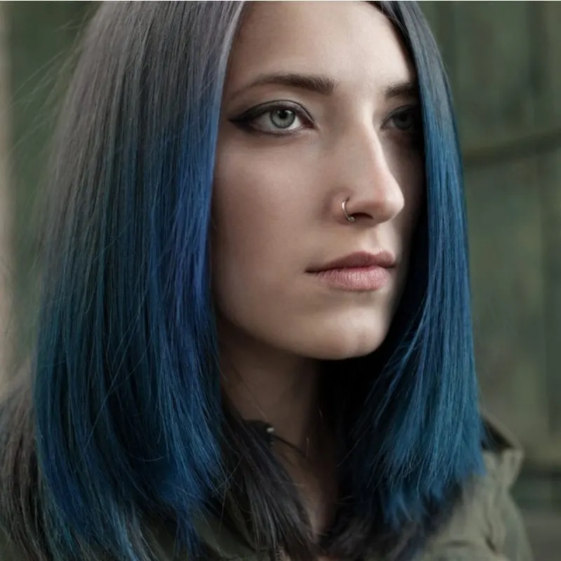 Woman with a nose ring and dark blue hair in a brown jacket stares straight ahead
