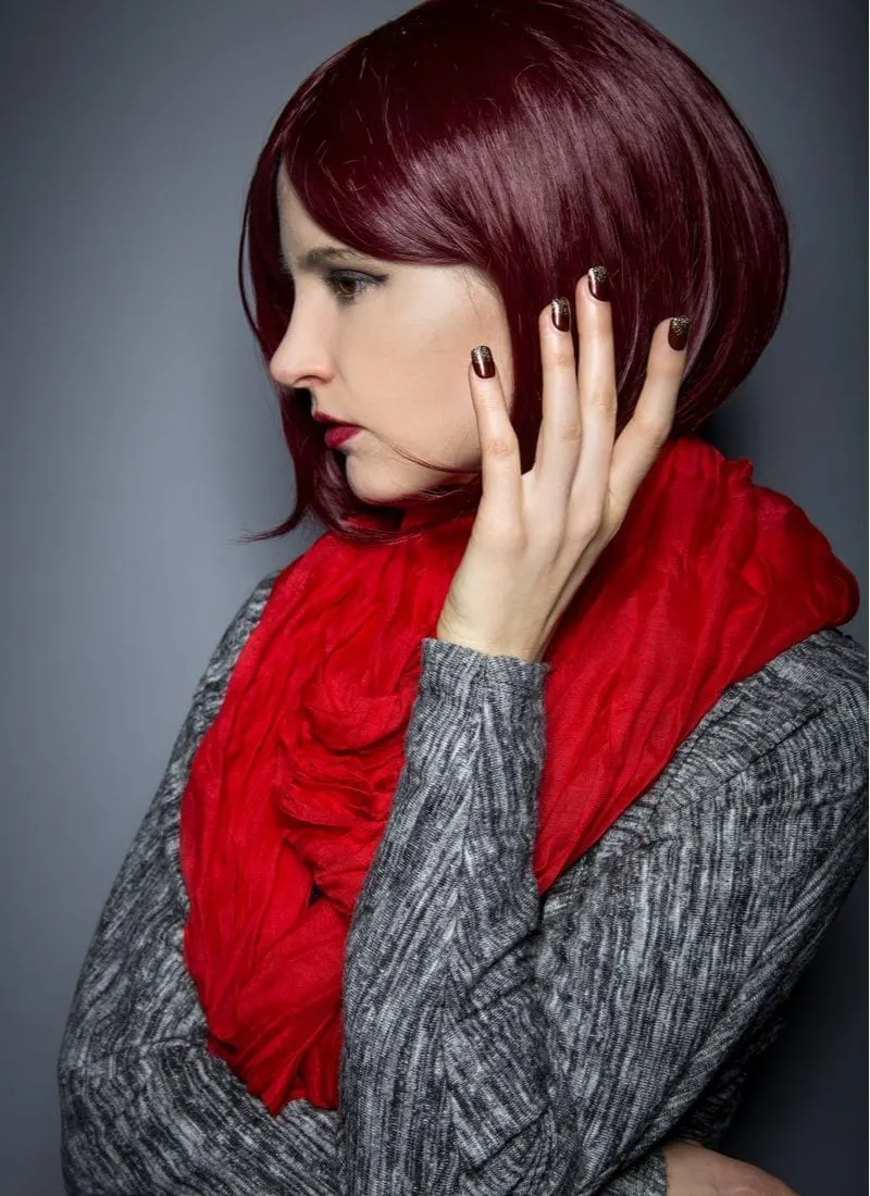 Woman in a grey heathered sweater holds her hand up to her face with burgundy color nails and hair and wearing a bright red scarf