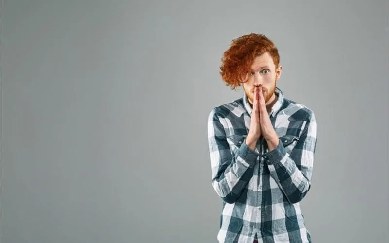 Portrait of a man with a red hairstyle holding his hands up to this mouth in a prayer motion and rocking a flannel button up shirt