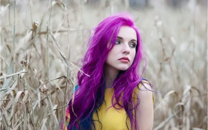 Woman with purple hair and wearing a yellow shirt looks up to her left while standing in a field of high grass