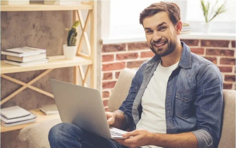 Man with a widows peak smiles at the camera and works on his laptop