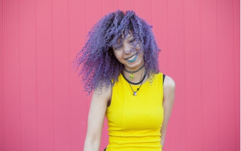 Woman standing in a yellow dress and wearing purple hair against a pink background