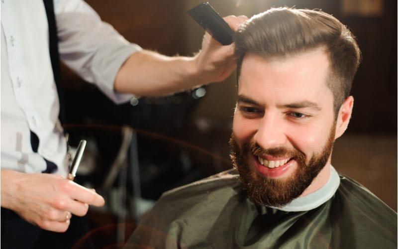 Guy in a barber chair wears a cape while his barber gives him an up and over tapered fade haircut
