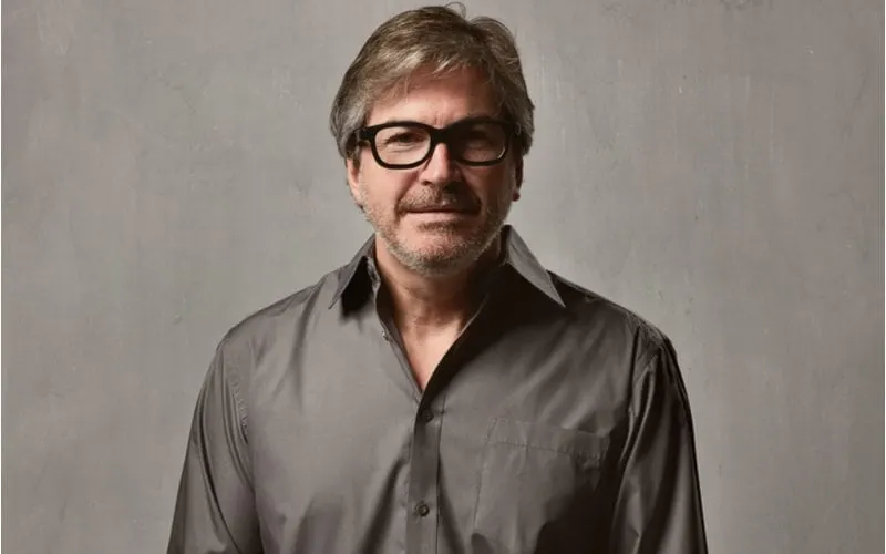 Man in a grey shirt that is shiny puts his hands in his pockets and eyes the camera through his thick-rimmed glasses
