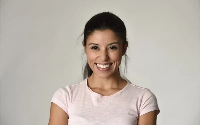 Woman with a heathered white v neck shirt smiles at the camera and wears a standard latina hairstyle