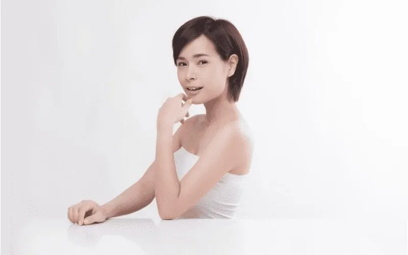 Asian short haired woman holds her chin and sits at an all-white desk against a white background