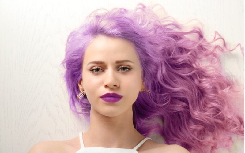 Woman with lilac hair lays on a light wood floor and wears purple lipstick