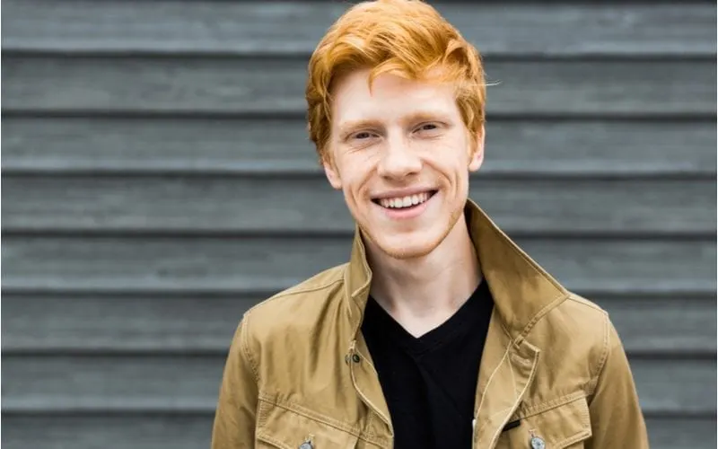 Young red headed man smiles and looks into the camera while standing outside of a barn looking building