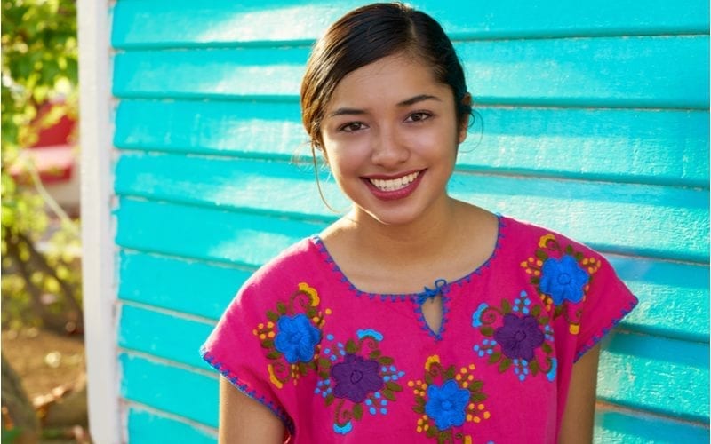 Young Mexican woman in a pink floral shirt stands in front of an aging blue sided building