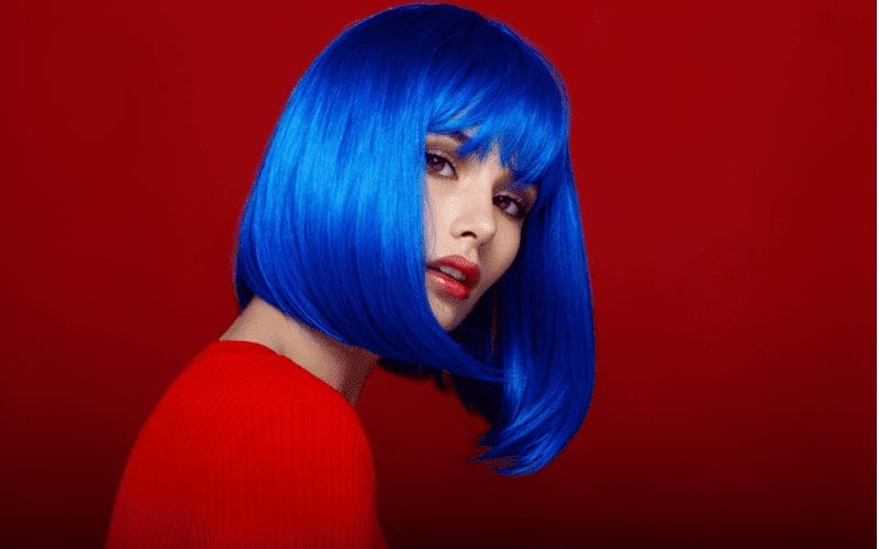 Young lady with a long blue bob haircut wears a red sweater and stands against a darker blue wall
