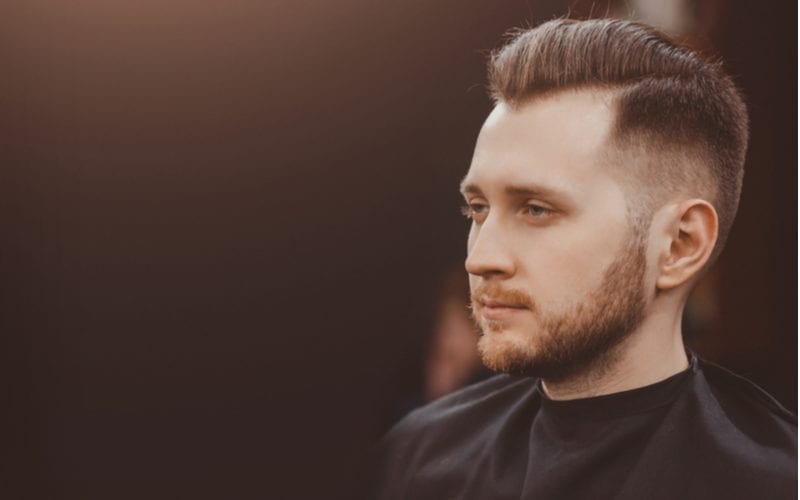 Man with a widows peak gets a haircut in a barber chair while wearing a cape