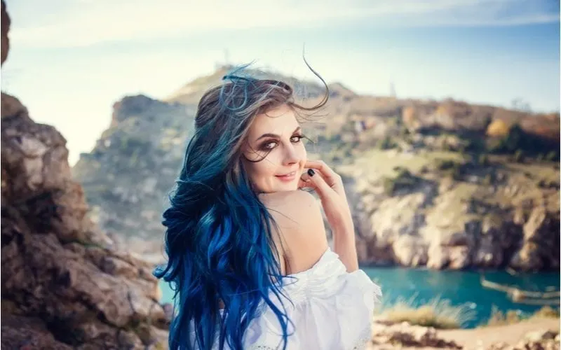 Woman standing with blue hair and looking back at the camera in front of lakes and cliffs