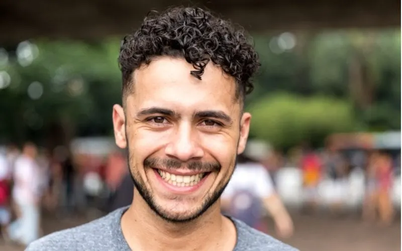 Brazilian man with a curly tight bowl cut and a thin beard and mustache stands outside smiling