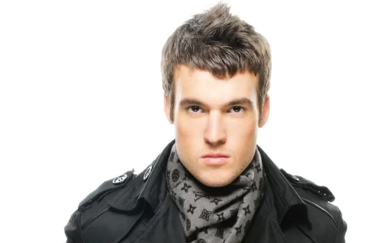 Man in a trenchcoat wears a haircut for guys with big foreheads and looks into the camera against a plain white background