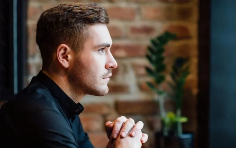 Guy with a mid-length top and faded sides haircut sits and looks out the window of a coffee shop with a brick wall to his left